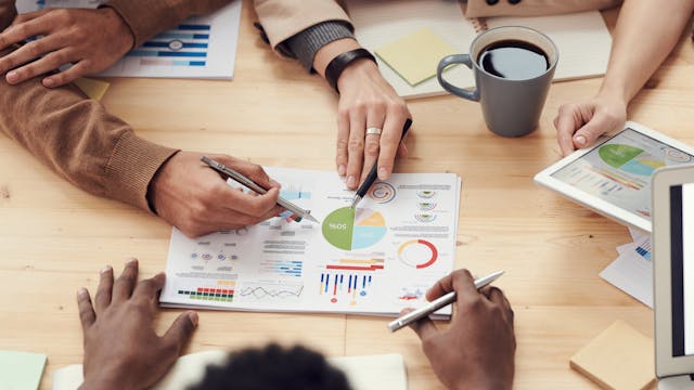 The picture shows hands of different races pointing to a document full of graphs. This represents seeking financial sustainability.